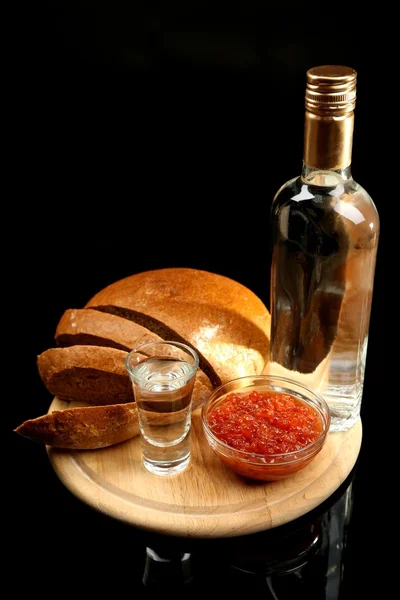 Láhev vodky, červený kaviár, čerstvý chléb na dřevěné desce, izolované na černém pozadí — Stockfoto