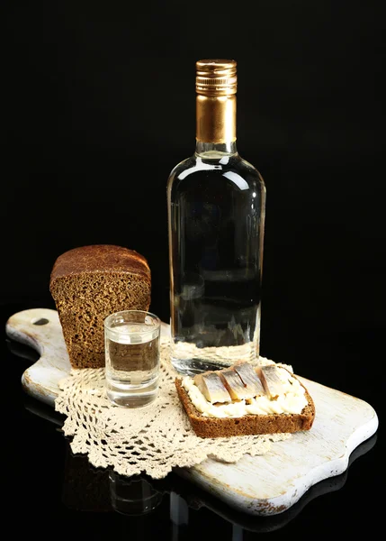 Láhev vodky, sendvič s solené ryby a brýle na dřevěné desce, izolované na černém — Stock fotografie