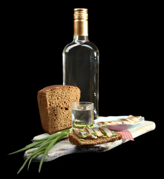 Composição com garrafa de vodka, sanduíche com peixe salgado, cebola verde e vidro sobre tábua de madeira, isolado em preto — Fotografia de Stock