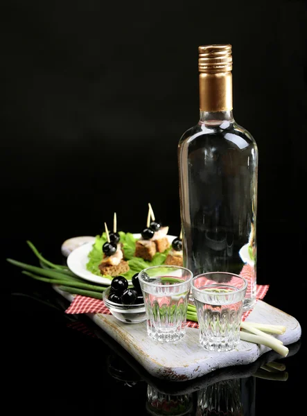 Склад з пляшкою горілки, закуски з солоною рибою, зелена цибуля і склянка на дерев'яній дошці, ізольовані на чорному — стокове фото