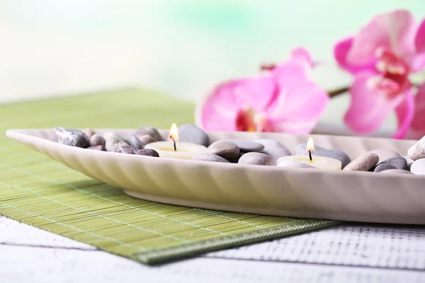 Spa stones and candle in decorative bowl, on wicker mat, on color wooden background — Stock Photo, Image