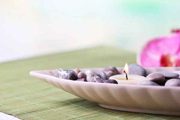 Spa stones and candle in decorative bowl, on wicker mat, on color wooden background — Stock Photo, Image