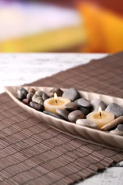 Pedras de spa e vela em tigela decorativa, no tapete de bambu, na mesa de madeira colorida, no fundo brilhante — Fotografia de Stock