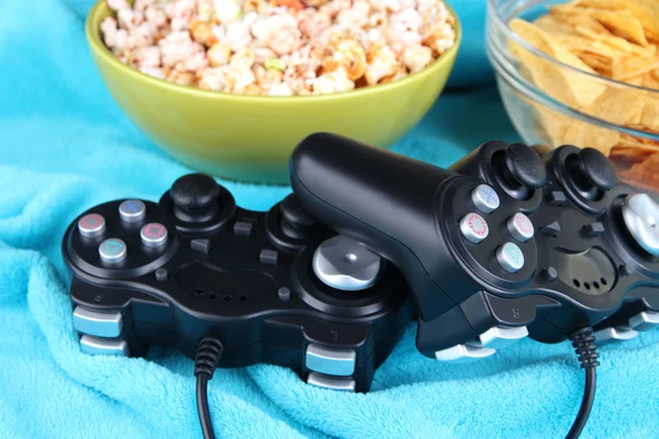 Black game controllers and bowl with snacks on color plaid background — Stock Photo, Image