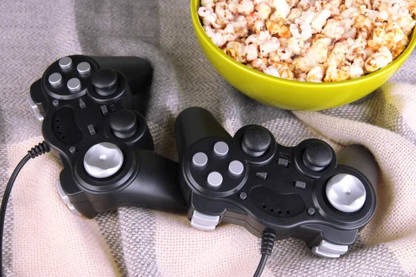 Black game controllers and bowl with pop corn on color plaid background — Stock Photo, Image