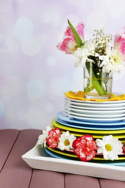 堆多彩陶瓷餐具和鲜花的托盘，木桌前，在明亮的背景上 — 图库照片