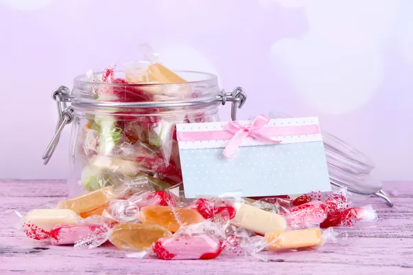 Sabrosos caramelos en frasco con tarjeta en la mesa sobre fondo brillante —  Fotos de Stock