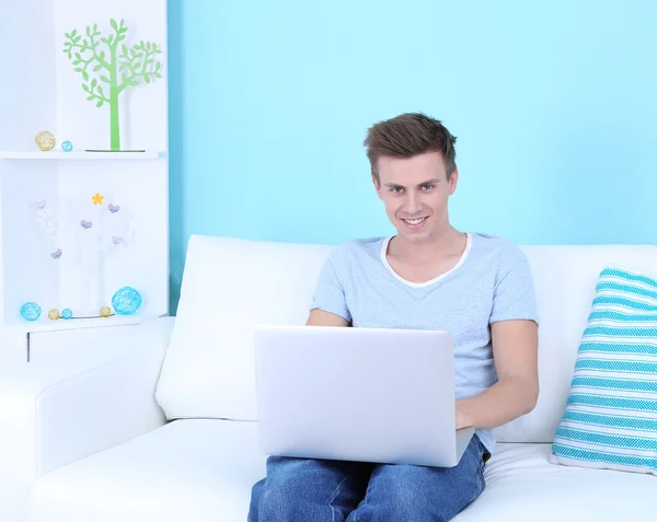 Guy sitting on sofa with laptop on blue background — Stock Photo, Image