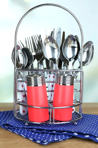 Kitchen cutlery in metal stand on wooden table on bright background — Stock Photo, Image