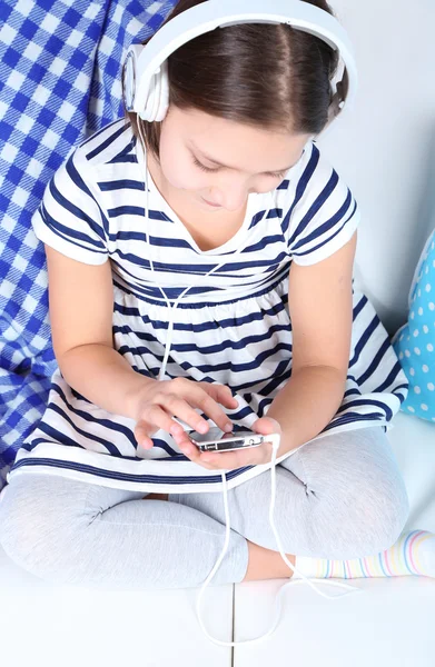 Beautiful little girl sitting on sofa  and listening to music, on home interior background — Stock Photo, Image