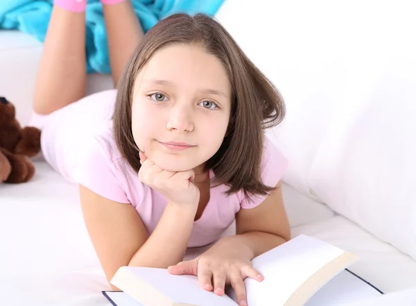 Bella bambina seduta sul divano con libro, su sfondo interno di casa — Foto Stock