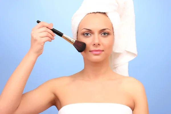 Красивая молодая женщина после душа с полотенцем на голове и кистью макияж в руке на синем фоне крупным планом — стоковое фото