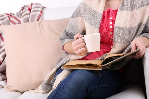 Γυναίκα κάθεται στον καναπέ του, διαβάζοντας το βιβλίο και καφέ ή τσάι, κινηματογράφηση σε πρώτο πλάνο — Φωτογραφία Αρχείου
