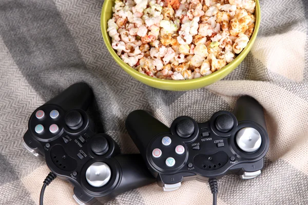 Black game controllers and bowl with pop corn on color plaid background — Stock Photo, Image