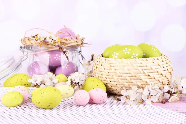 Composition with Easter eggs in glass jar and wicker basket, and blooming branches on light background — Stock Photo, Image