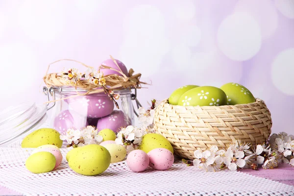 Composition with Easter eggs in glass jar and wicker basket, and blooming branches on light background — Stock Photo, Image