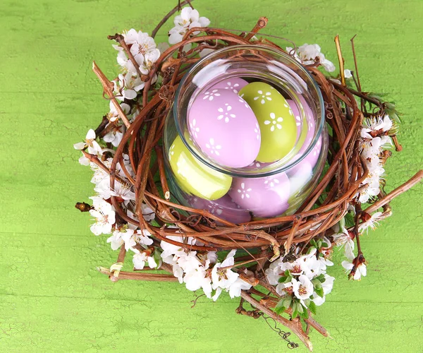 Composition with Easter eggs and blooming branches in glass jar and decorative nest, on  color wooden background — Stock Photo, Image