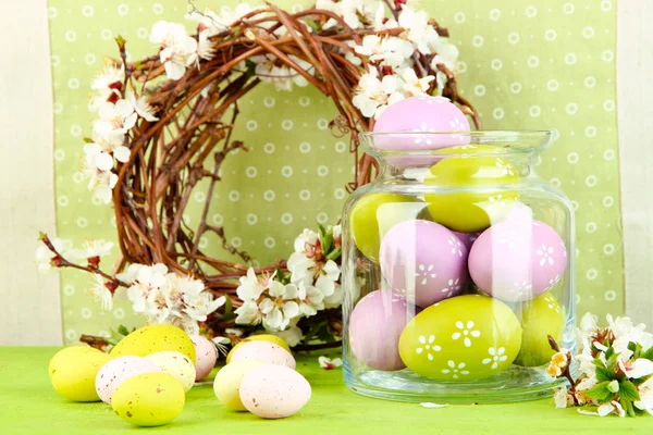 Composition with Easter eggs in glass jar and decorative wreath with blooming branches on light background — Stock Photo, Image