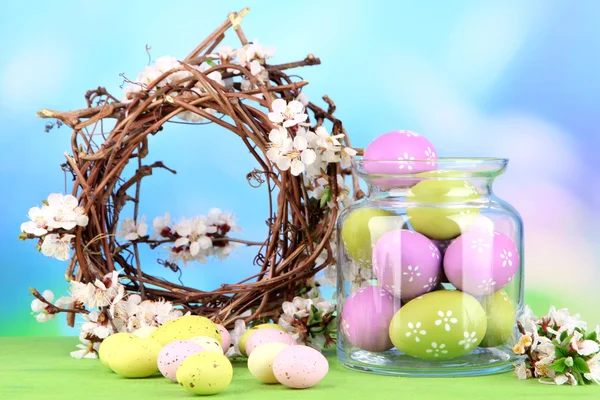 Sammansättning med påskägg i glasburk och dekorativ krans med blommande grenar på ljus bakgrund — Stockfoto