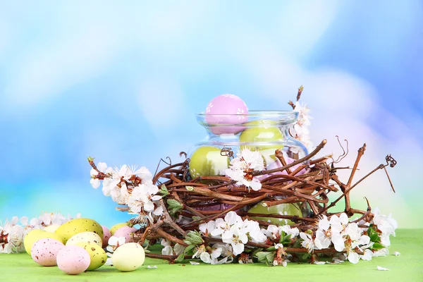 Composition with Easter eggs and blooming branches in glass jar and decorative nest, on color wooden table, on bright background — Stock Photo, Image