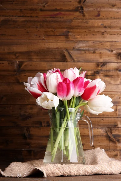 Krásné tulipány ve skleněné konvici na dřevěné pozadí — Stock fotografie