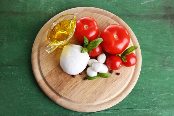 Composição com saborosas bolas de queijo mussarela, manjericão e tomates vermelhos, azeite na tábua de corte, na cor de fundo de madeira — Fotografia de Stock