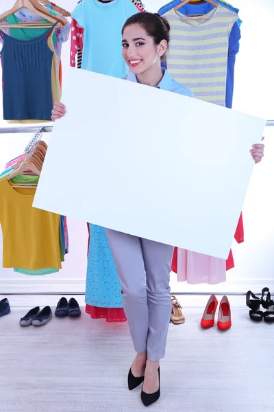 Hermosa joven sosteniendo cartel en blanco cerca de rack con perchas — Foto de Stock