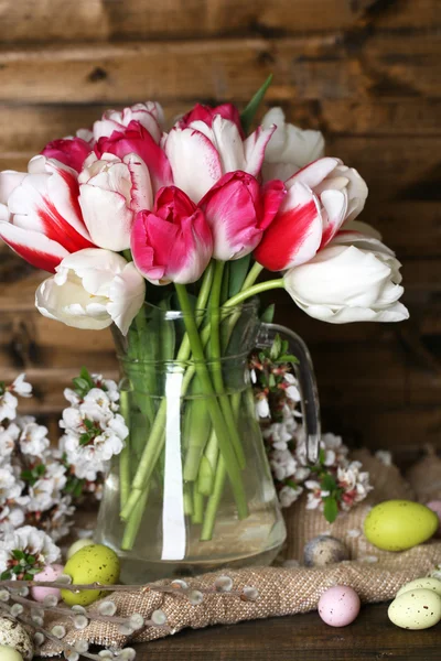 Composition with Easter eggs and beautiful tulips in glass jug on wooden background — Stock Photo, Image