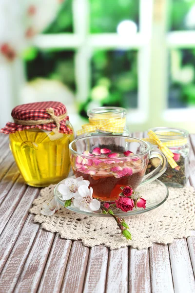 Ассортимент трав, чая и меда в стеклянных банках и чашка горячего напитка на деревянном столе, на ярком фоне — стоковое фото