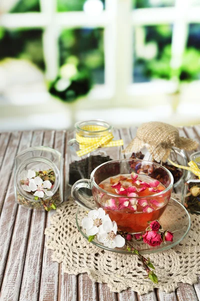 Ассортимент трав, чай в стеклянных банках и горячий напиток в чашке на деревянном столе, на ярком фоне — стоковое фото