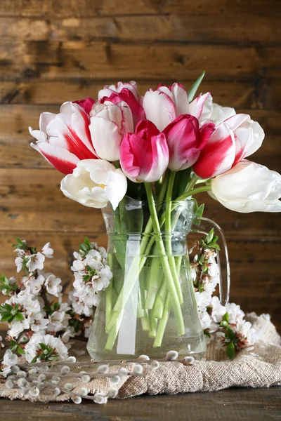 Krásné tulipány ve skleněné konvici na dřevěné pozadí — Stock fotografie