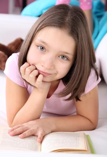 Hermosa niña sentada en el sofá con libro, en el fondo interior de casa — Foto de Stock