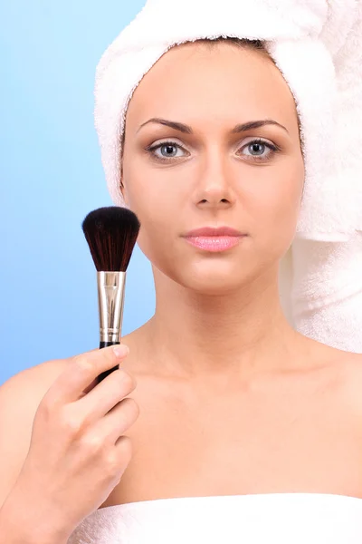 Hermosa joven después de la ducha con una toalla en la cabeza y un maquillaje de cepillo en la mano sobre un fondo azul — Foto de Stock