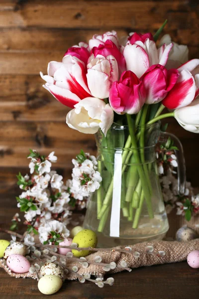 Composizione con uova di Pasqua e bellissimi tulipani in brocca di vetro su fondo di legno — Foto Stock