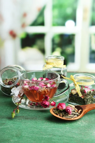 Assortimento di erbe aromatiche, tè in barattoli di vetro e bevanda calda in tazza su tavolo di legno, su sfondo luminoso — Foto Stock