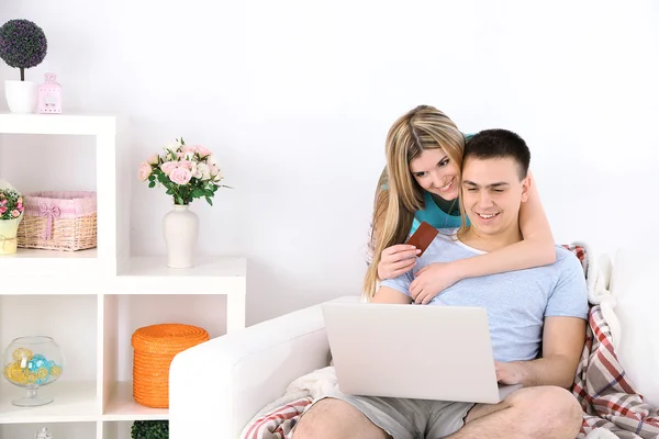 Coppia amorevole seduta con computer portatile e carta di credito sul divano, su sfondo interno di casa — Foto Stock