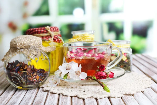 Assortiment van kruiden, thee en honing in glazen potten en kopje warme drank op houten tafel, op lichte achtergrond — Stockfoto