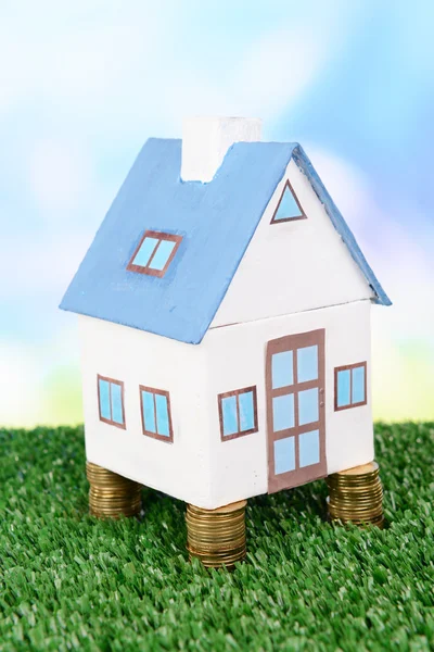 Дом стоит на куче монет на траве на ярком фоне — стоковое фото