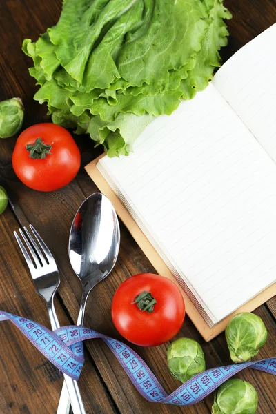 Cutlery tied with measuring tape and book with vegetables on wooden background — Stock Photo, Image