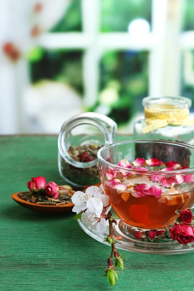 Ассортимент трав, чай в стеклянных банках и горячий напиток в чашке на деревянном столе, на ярком фоне — стоковое фото