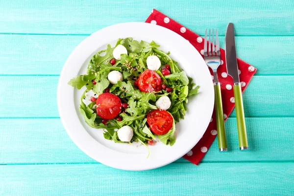 Зеленый салат из рукколы, помидоров, сыра моцарелла шарики и кунжута на тарелке, на цветном деревянном фоне — стоковое фото
