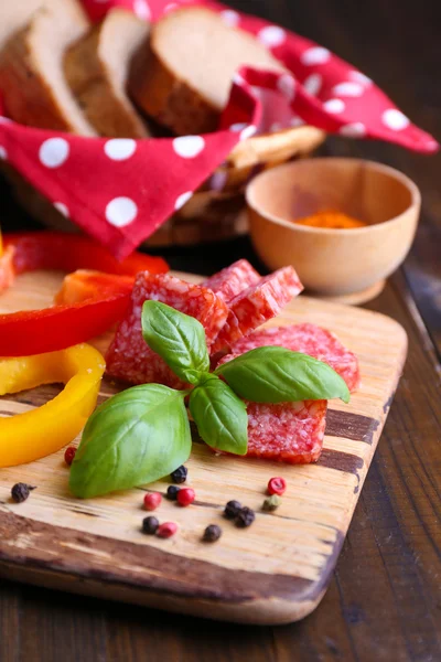 Склад з ножем, сосискою салямі, нарізаним хлібом і перцем на обробній дошці, на дерев'яному фоні — стокове фото