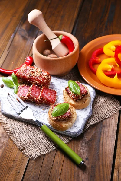 Склад з ножем, смачні бутерброди з салямі ковбаса, листя базиліка на обробній дошці, на дерев'яному фоні — стокове фото