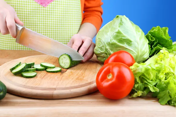 Manos femeninas cortando pepino sobre tabla de madera, primer plano, sobre fondo azul — Foto de Stock