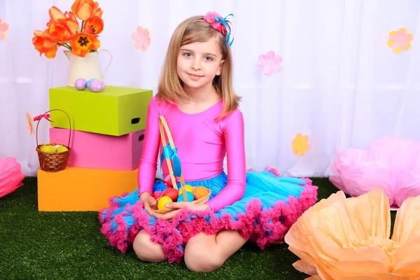 Hermosa niña pequeña en falda pequeña que sostiene la cesta con huevos de colores sobre fondo decorativo — Foto de Stock