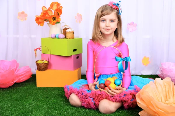 Красива маленька дівчинка в дрібній спідниці тримає кошик з барвистими яйцями на декоративному фоні — стокове фото