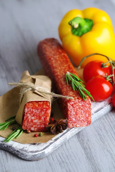 Tasty salami sausage, vegetables and spices on paper on wooden background — Stock Photo, Image