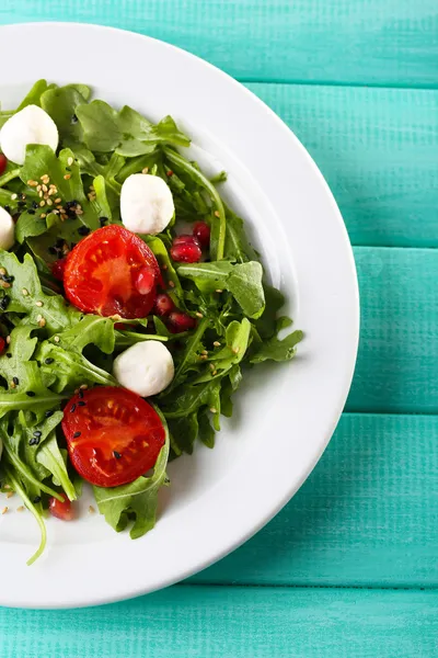 Grüner Salat mit Rucola, Tomaten, Käse-Mozzarella-Bällchen und Sesam auf Teller, auf farbigem Holzhintergrund — Stockfoto