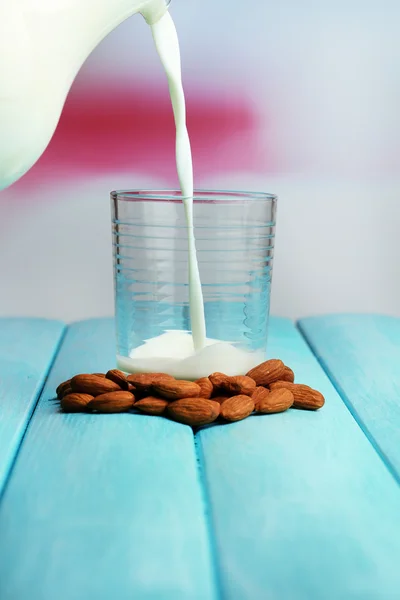 Αμύγδαλο γάλα χύνεται σε γυαλί, σε χρώμα ξύλινο τραπέζι, επάνω ελαφρύς υπόβαθρο — Φωτογραφία Αρχείου