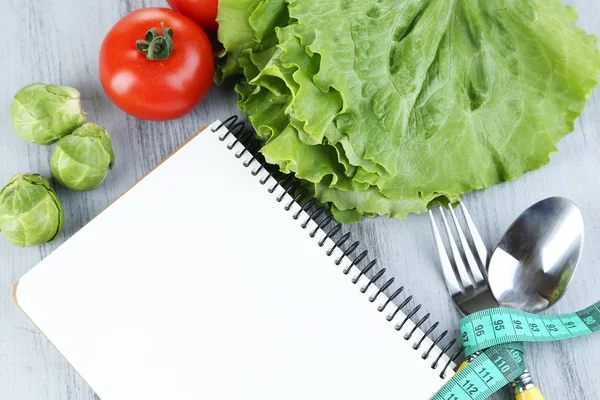 Cutlery tied with measuring tape and notebook with vegetables on wooden background — Stock Photo, Image